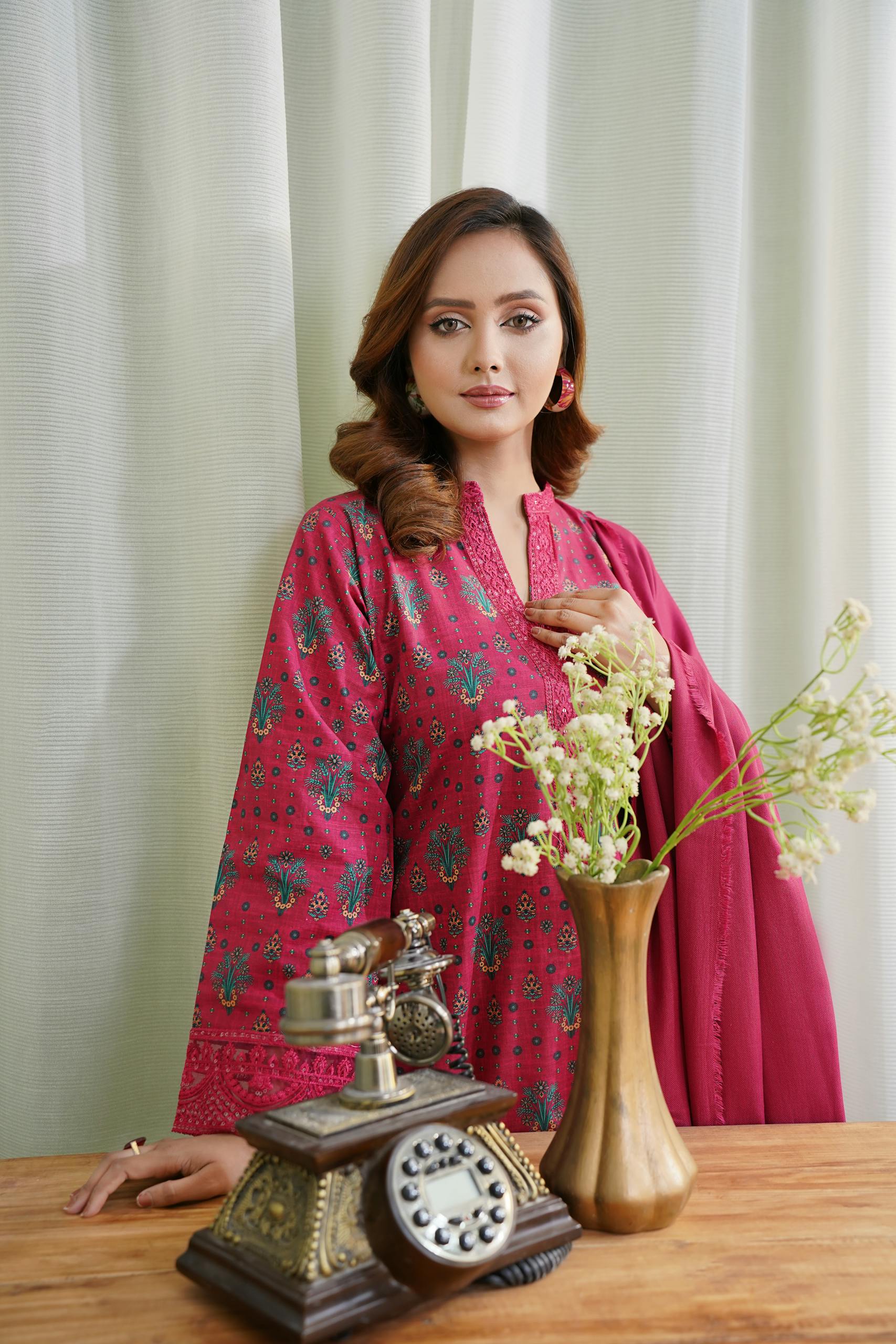 Woman in a vibrant pink Pakistani dress stands beside vintage telephone and vase in Lahore, Pakistan.